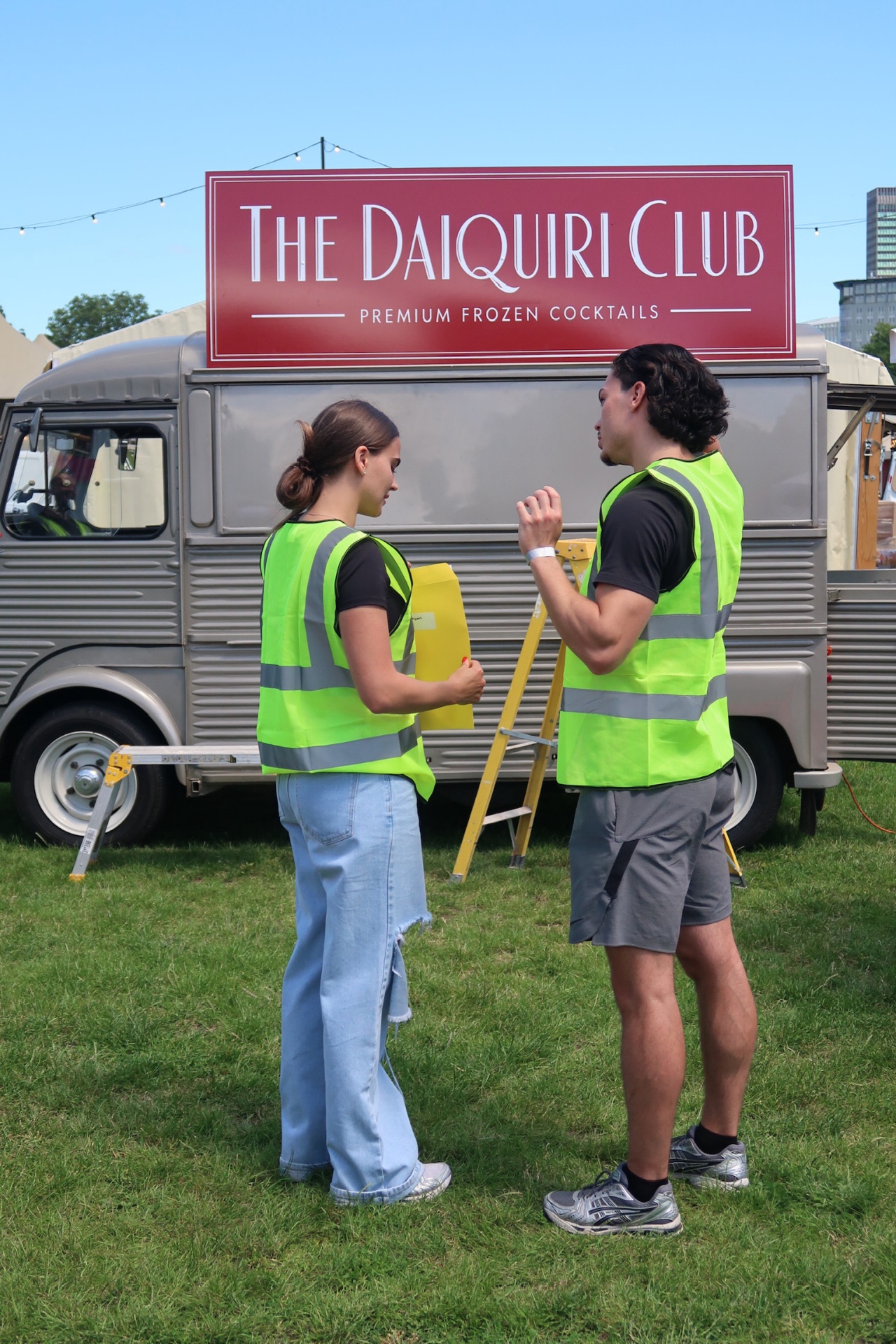 Ruby and Noah in hi-vis vests in front of the van sign